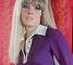 Wendy Richard, wearing a long wig, wearing a purple dress, sitting on a barstool